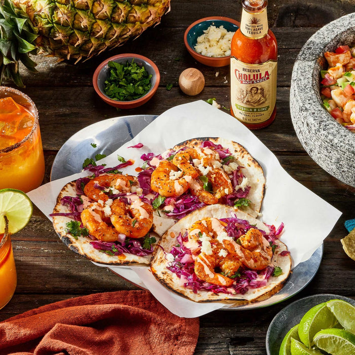 Two shrimp tacos with red cabbage, cilantro, and a white sauce on a white paper on a wooden table with a bottle of Reserva Cholula Hot Sauce.