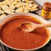 A white bowl of Cholula Original Medium Salsa with a wooden spoon in it, next to a baking sheet of tortilla chips.