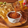 A jar of Cholula Original Medium Salsa is being poured into a white bowl with tortilla chips on a wooden tray.