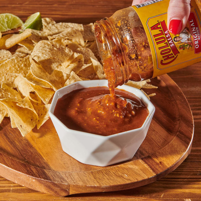 A jar of Cholula Original Medium Salsa is being poured into a white bowl with tortilla chips on a wooden tray.