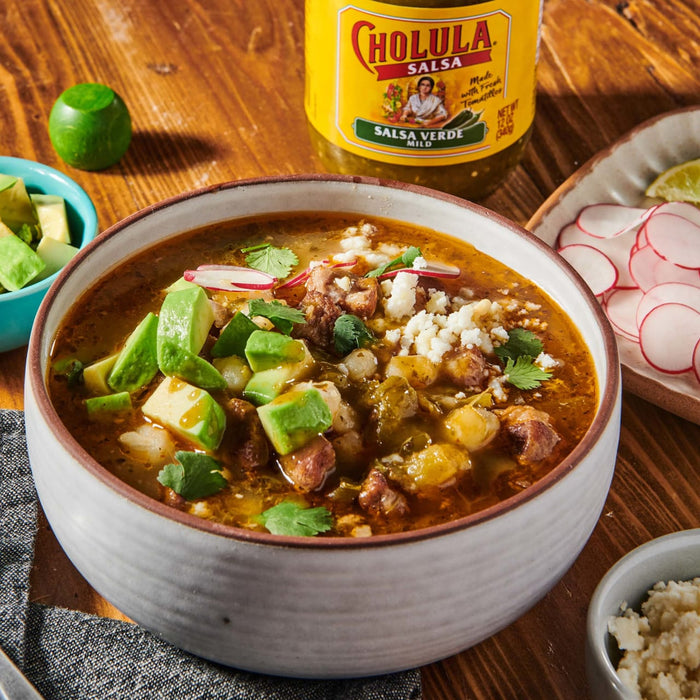 A bowl of Mexican soup with avocado, radish, and cilantro, topped with crumbled cheese, with a bottle of Cholula Salsa Verde Mild in the background.