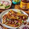 Two tacos filled with fish, avocado, red onion, and corn are topped with Cholula Smoky Chipotle Hot Salsa.