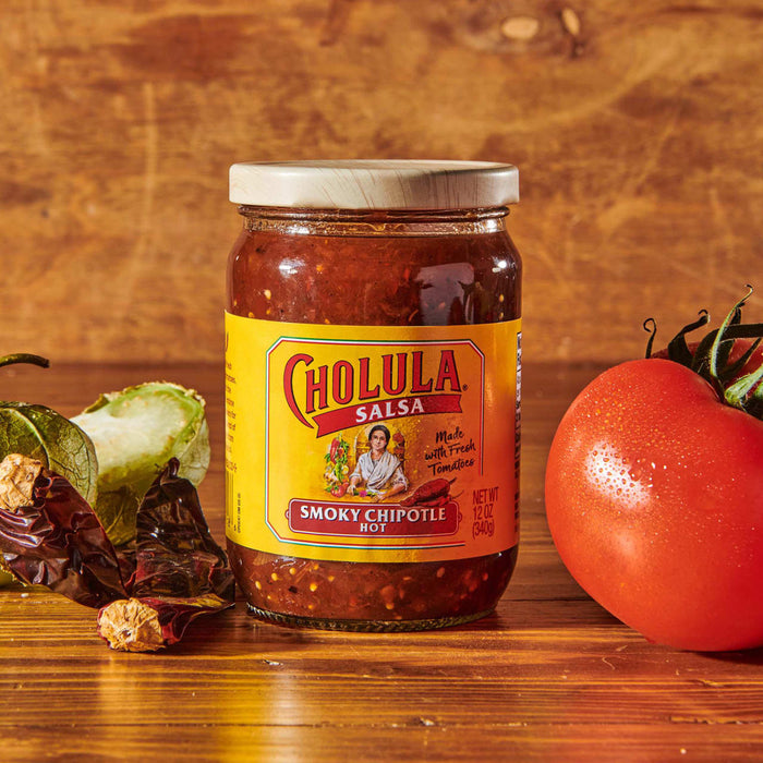 A jar of Cholula Smoky Chipotle Hot Salsa sits on a wooden table with a tomato and dried peppers.