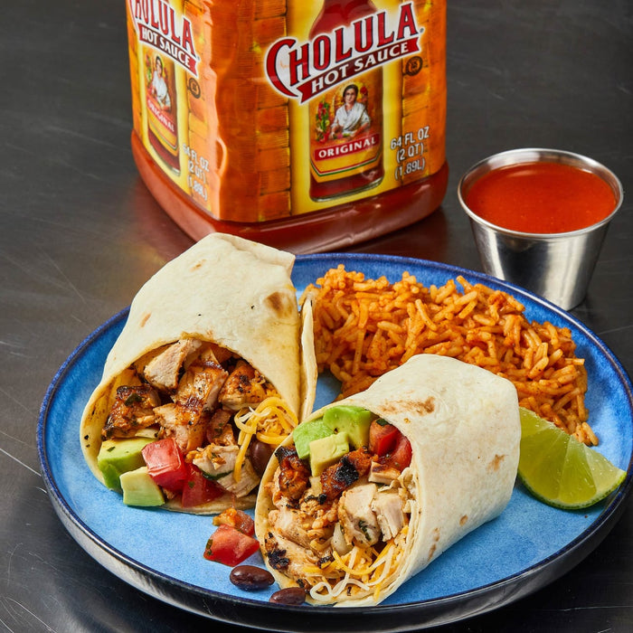 Two burritos with chicken, avocado, tomato, and cheese on a blue plate with rice and a lime wedge, next to a bottle of Cholula Hot Sauce.