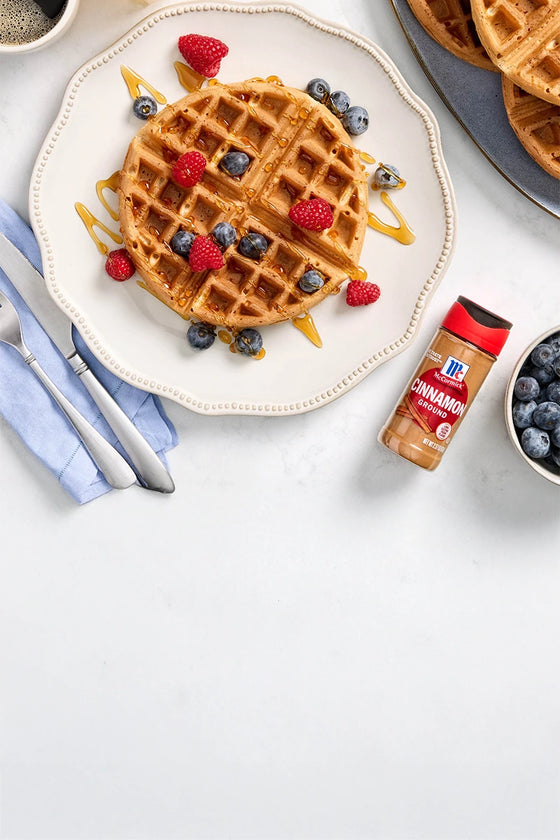 Waffle with berries on a plate, surrounded by coffee, syrup, and cinnamon.