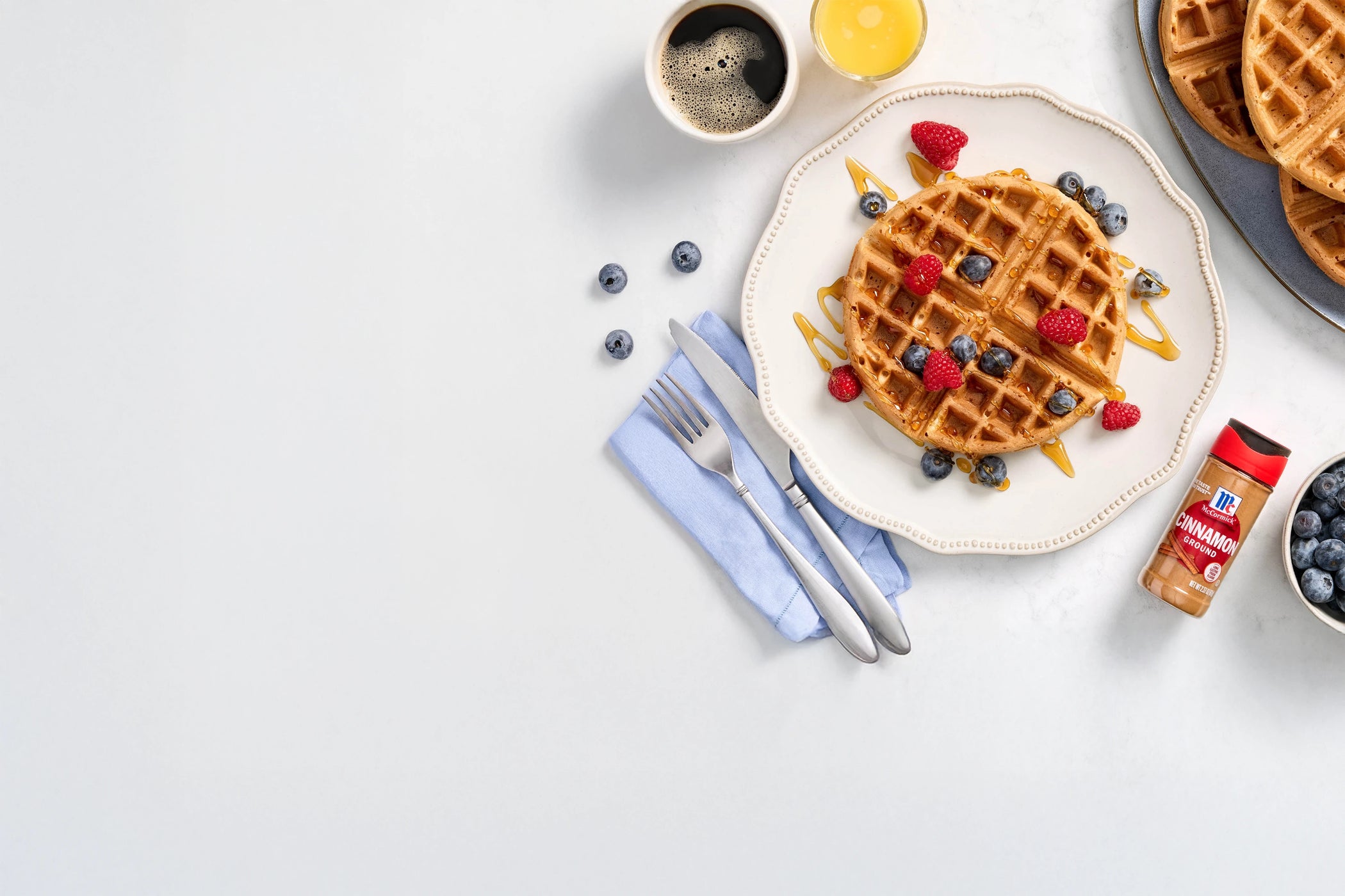 Waffle with berries on a plate, surrounded by coffee, syrup, and cinnamon.