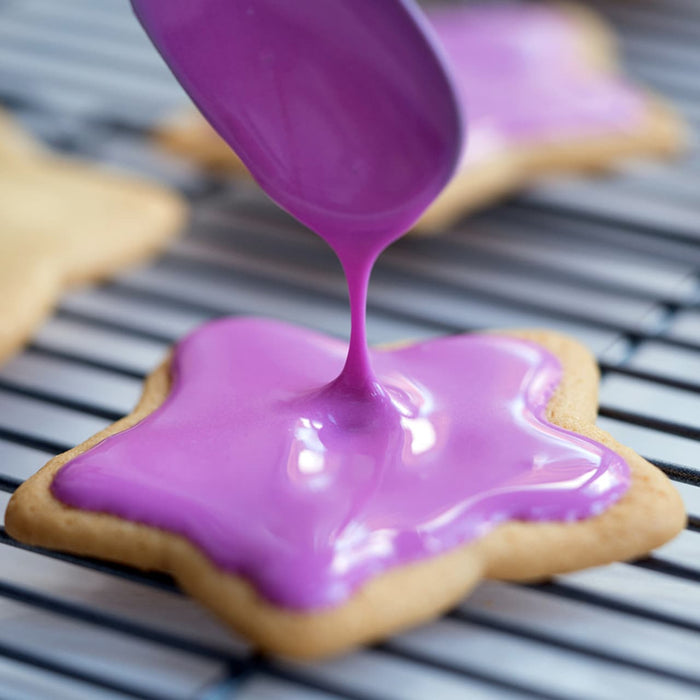 A star-shaped cookie is being decorated with purple icing on a wire rack.