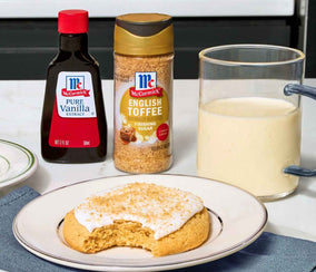 Baked cookie with cream on a plate, next to McCormick vanilla extract, English toffee finishing sugar, and a glass of milk.