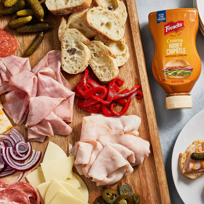 A wooden cutting board with a variety of meats, cheeses, and vegetables, including a jar of French's Creamy Honey Chipotle Mustard Spread.