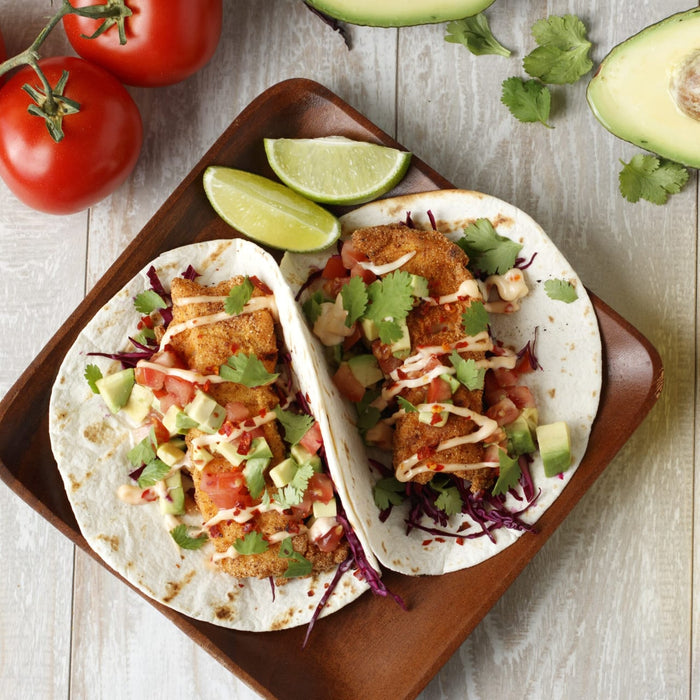 Two fish tacos with avocado, tomato, and lime on a wooden plate.