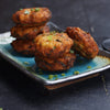 A stack of golden-brown fried patties, garnished with chopped green onions, sits on a blue and brown square plate against a dark background.