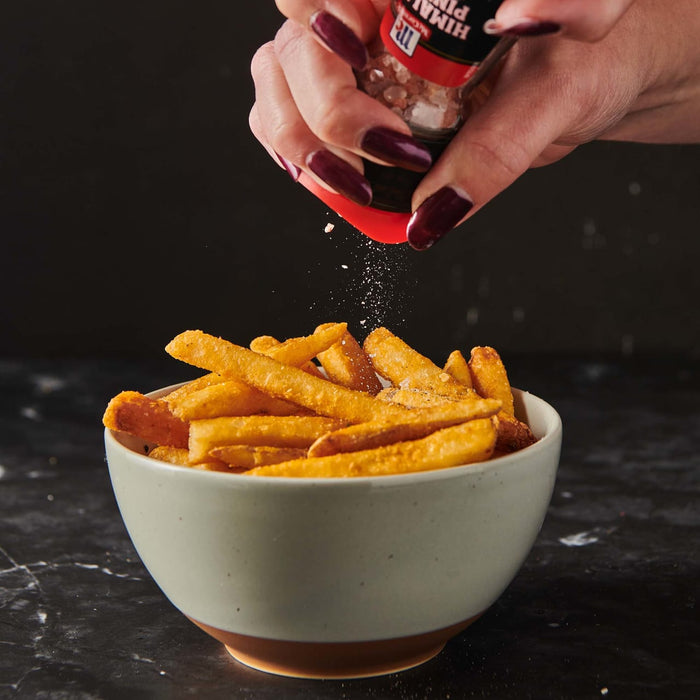 A hand sprinkles pink Himalayan salt from a McCormick shaker onto a bowl of french fries.