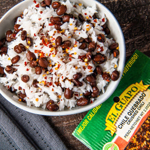 A bowl of white rice with black beans and red pepper flakes, next to a package of El Guapo Crushed Chili.