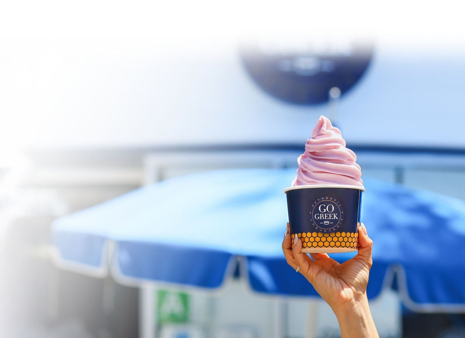 go greek cup of black currant greek yogurt being held in front of storefront with a blue umbrella out front