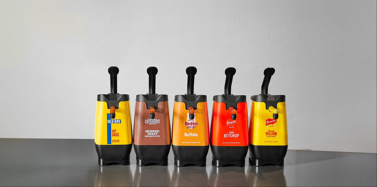 Five hot dog condiment squeeze bottles lined up on a dark reflective surface against a light gray background. The bottles are yellow, brown, orange, red, and yellow, with black tops and visible labels for brands like KFC, Chick-fil-A, Taco Bell, and Wendy's.