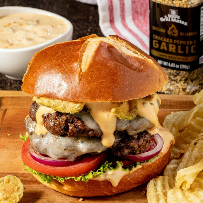 A double cheeseburger with fried pickles, lettuce, tomato, and red onion on a pretzel bun, with a side of potato chips and a jar of Grill Mates Cracked Pepper &amp; Garlic seasoning.