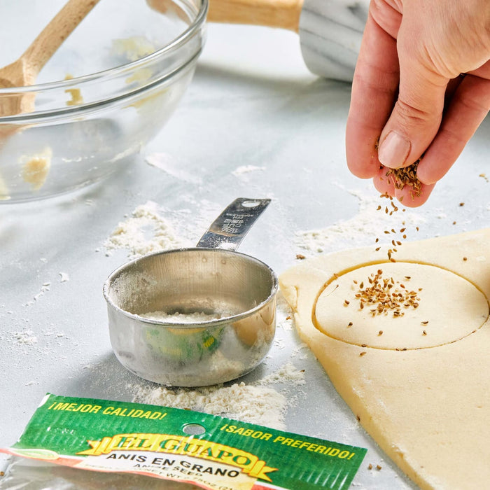 A hand sprinkles anise seeds onto a circle of dough, with a measuring cup and a bag of El Guapo anise seeds in the background.