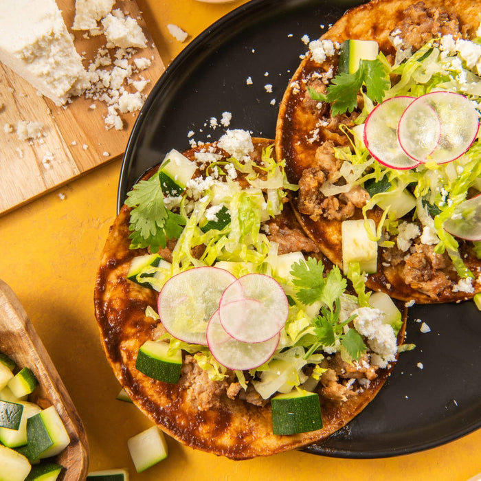 Two tostadas topped with meat, lettuce, zucchini, radish, cilantro, and crumbled cheese.