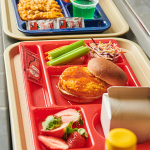A red school lunch tray features a crispy chicken sandwich with a packet of Frank's RedHot sauce alongside fresh strawberries, celery sticks, and coleslaw.