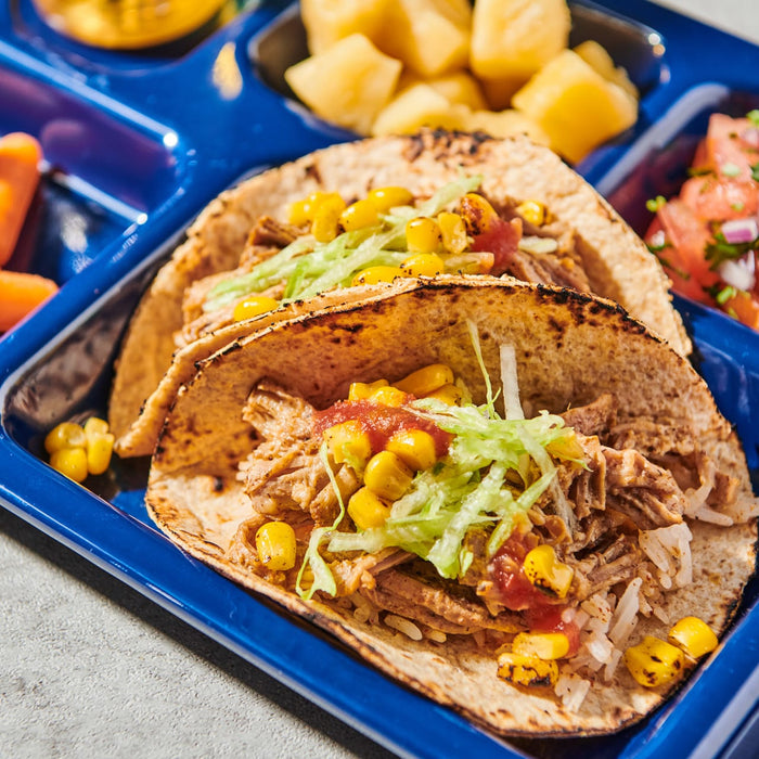 Two carnitas tacos filled with rice, shredded pork, lettuce, and charred corn are served on a blue tray alongside fresh pineapple and pico de gallo.