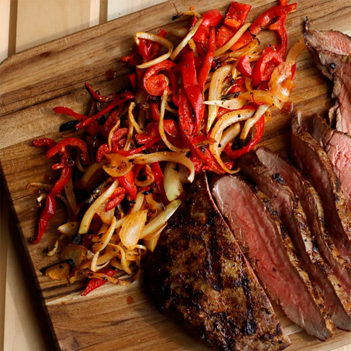 A grilled steak, sliced to show its medium-rare interior, is served on a wooden cutting board next to a pile of sautéed red bell peppers and onions, a meal enhanced by Mccormick Grill Mates Chipotle &amp; Roasted Garlic Seasoning.
