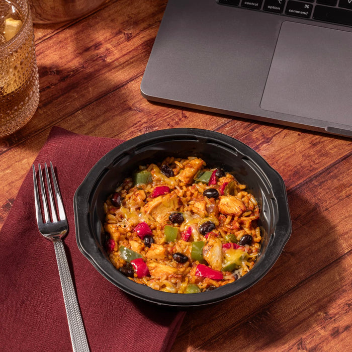 A black bowl of chicken and rice with black beans, peppers, and cheese sits on a wooden table next to a laptop and a glass of iced tea.