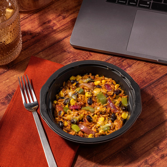 A bowl of rice with black beans, corn, peppers, and cheese on a wooden table with a fork and a glass of iced tea.