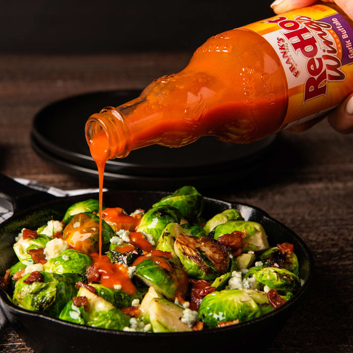 A bottle of Franks RedHot Garlic Buffalo Wing Sauce is being poured over a bowl of brussels sprouts, bacon, and blue cheese.