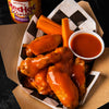A close-up of Franks RedHot Wings in a takeout container with carrots and a side of sauce.