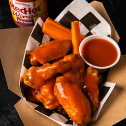 A close-up of Franks RedHot Wings in a takeout container with carrots and a side of sauce.