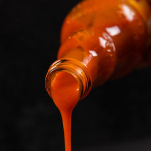 A bottle of Franks Red Hot Garlic Buffalo Wing Hot Sauce Regular 12 Oz is tilted, with the vibrant orange-red sauce pouring out of its neck against a dark background.