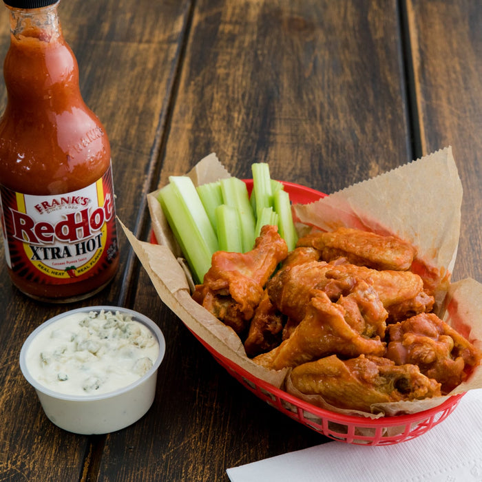 A bottle of Franks RedHot sauce, a basket of buffalo wings, celery sticks, and a bowl of blue cheese dressing.