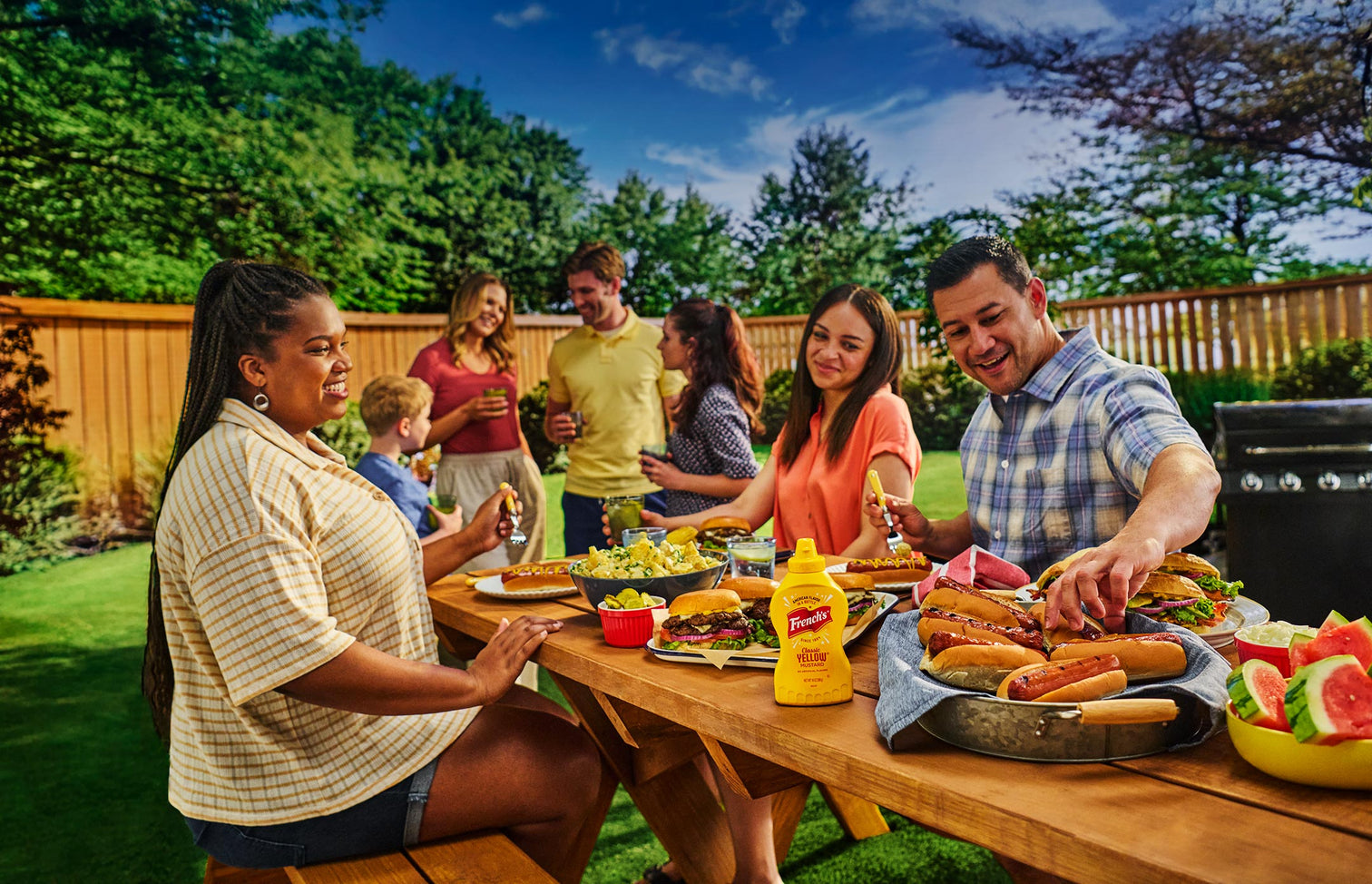friends and family sitting on a picnic bench enjoying outdoor grilling with hotdogs, burgers, potato salad, watermelon on a sunny day