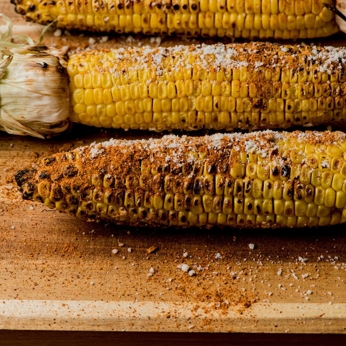 Three ears of corn are grilled and seasoned with salt and pepper on a wooden cutting board.