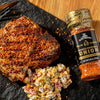 A close-up of a grilled pork chop with a side of corn salad and a jar of McCormick Grill Mates Smoked Paprika &amp; Onion seasoning.