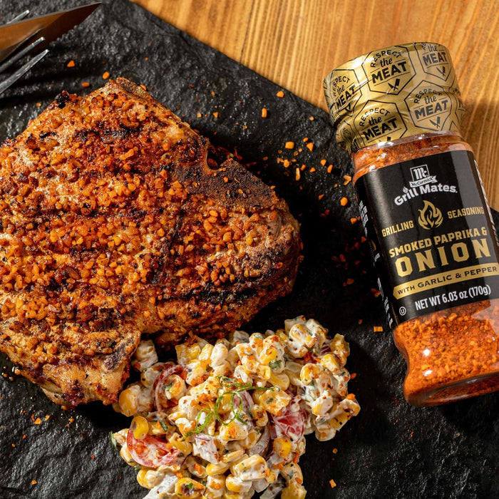 A close-up of a grilled pork chop with a side of corn salad and a jar of McCormick Grill Mates Smoked Paprika &amp; Onion seasoning.