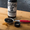 A bottle of Stubbs Hickory Liquid Smoke sits on a wooden cutting board with a measuring spoon and a lid.