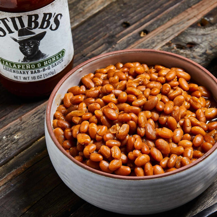 A bowl of baked beans sits on a wooden table next to a bottle of Stubbs BBQ sauce.