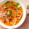  bowl of spicy noodle soup with lime, cilantro, and red onion on a pink background