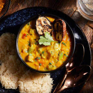 A vibrant bowl of vegetable curry featuring okra and grilled eggplant is served alongside a portion of fluffy white rice.