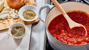 Cooking scene with tomato sauce in a pot, wooden spoon, and ingredients on a wooden board.