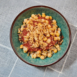 A vibrant bowl of koshary featuring layers of rice, lentils, and pasta topped with a rich tomato sauce, chickpeas, and crispy fried onions.