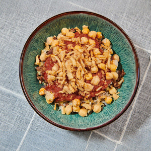 A vibrant bowl of koshary featuring layers of rice, lentils, and pasta topped with a rich tomato sauce, chickpeas, and crispy fried onions.