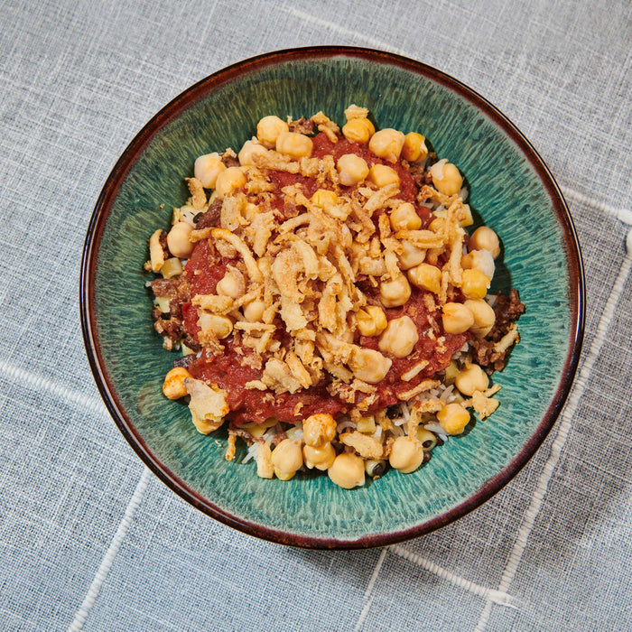 A vibrant bowl of koshary featuring layers of rice, lentils, and pasta topped with a rich tomato sauce, chickpeas, and crispy fried onions.