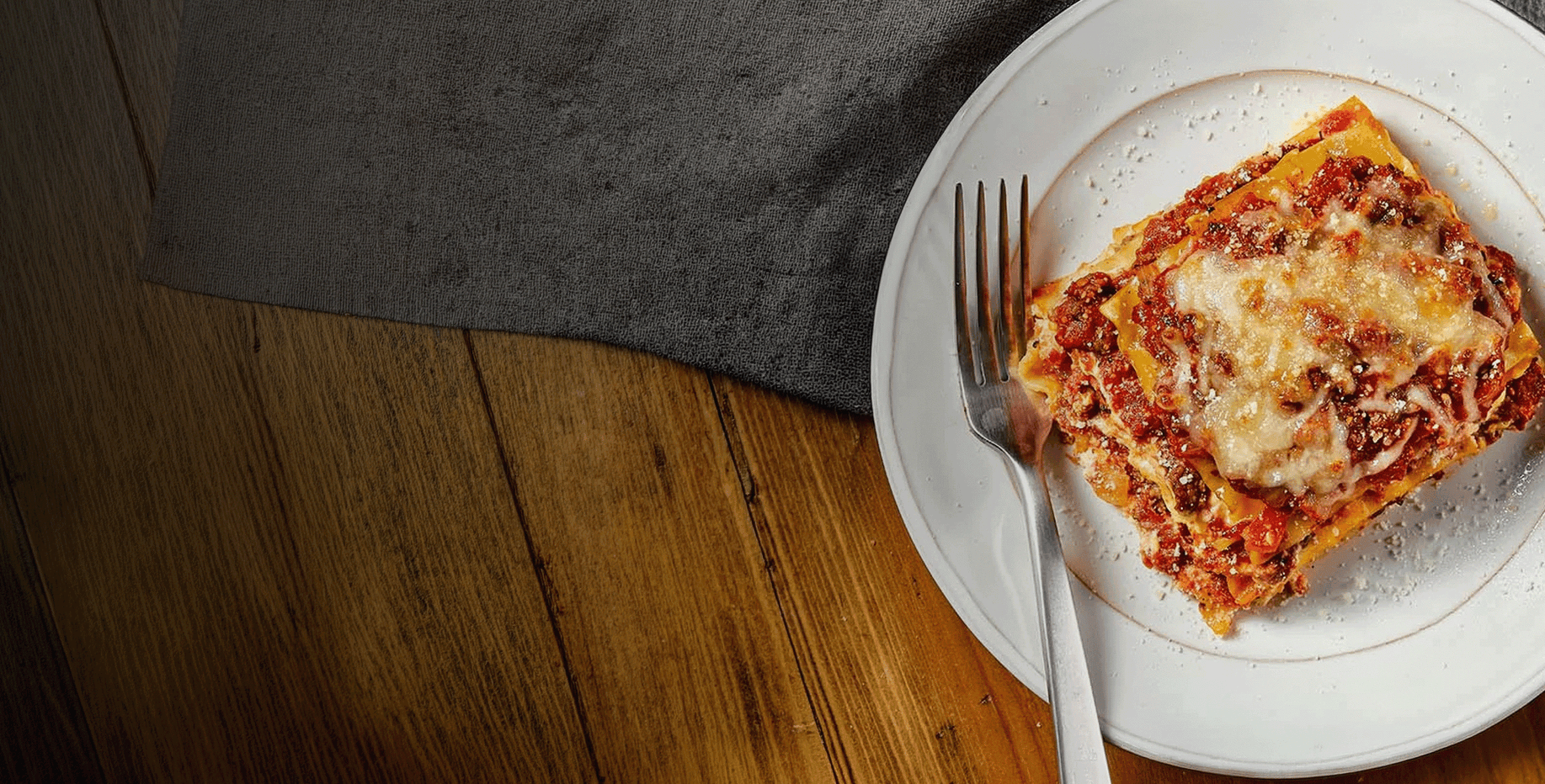 Slice of lasagna on a white plate with a fork, placed on a wooden table.