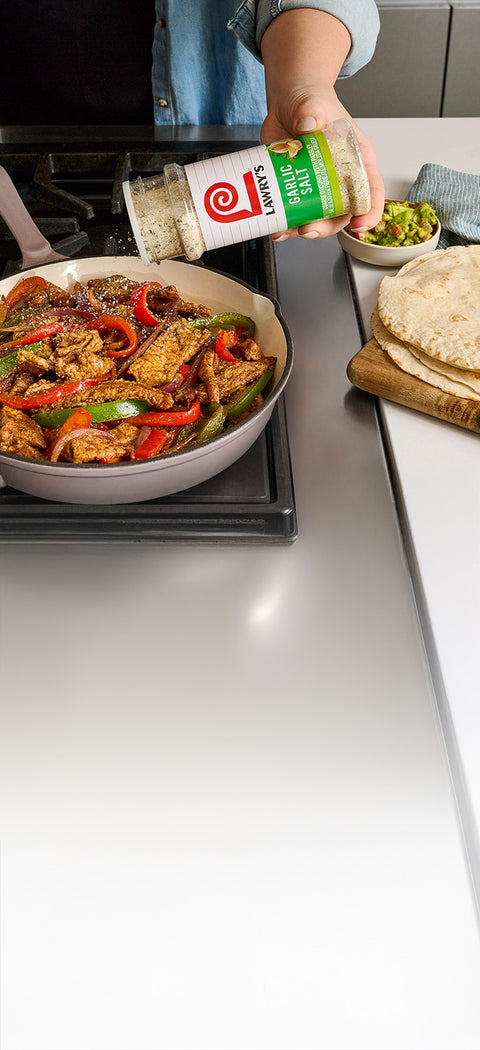 A person seasons fajitas in a pan with Lawry's Garlic Salt.