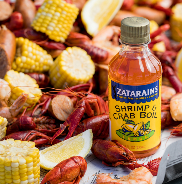 A bottle of Zatarains Shrimp &amp; Crab Boil Lemon seasoning sits on a table with crawfish, corn, and lemons.