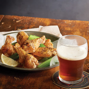 A plate of crispy chicken wings seasoned with black pepper and served with celery sticks and a lemon wedge next to a glass of amber beer.