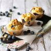 Three golden-brown blueberry muffins, likely enhanced with Mccormick Lemon Extract for a bright flavor, are displayed on a rustic wooden surface with scattered fresh blueberries.