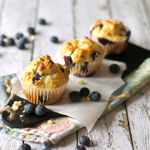 Three golden-brown blueberry muffins, likely enhanced with Mccormick Lemon Extract for a bright flavor, are displayed on a rustic wooden surface with scattered fresh blueberries.